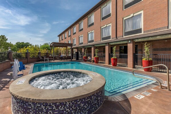 A swimming pool with a hot tub, surrounded by a brick building with large windows and outdoor seating. - Best Western Plus DFW Airport Suites (Irving)