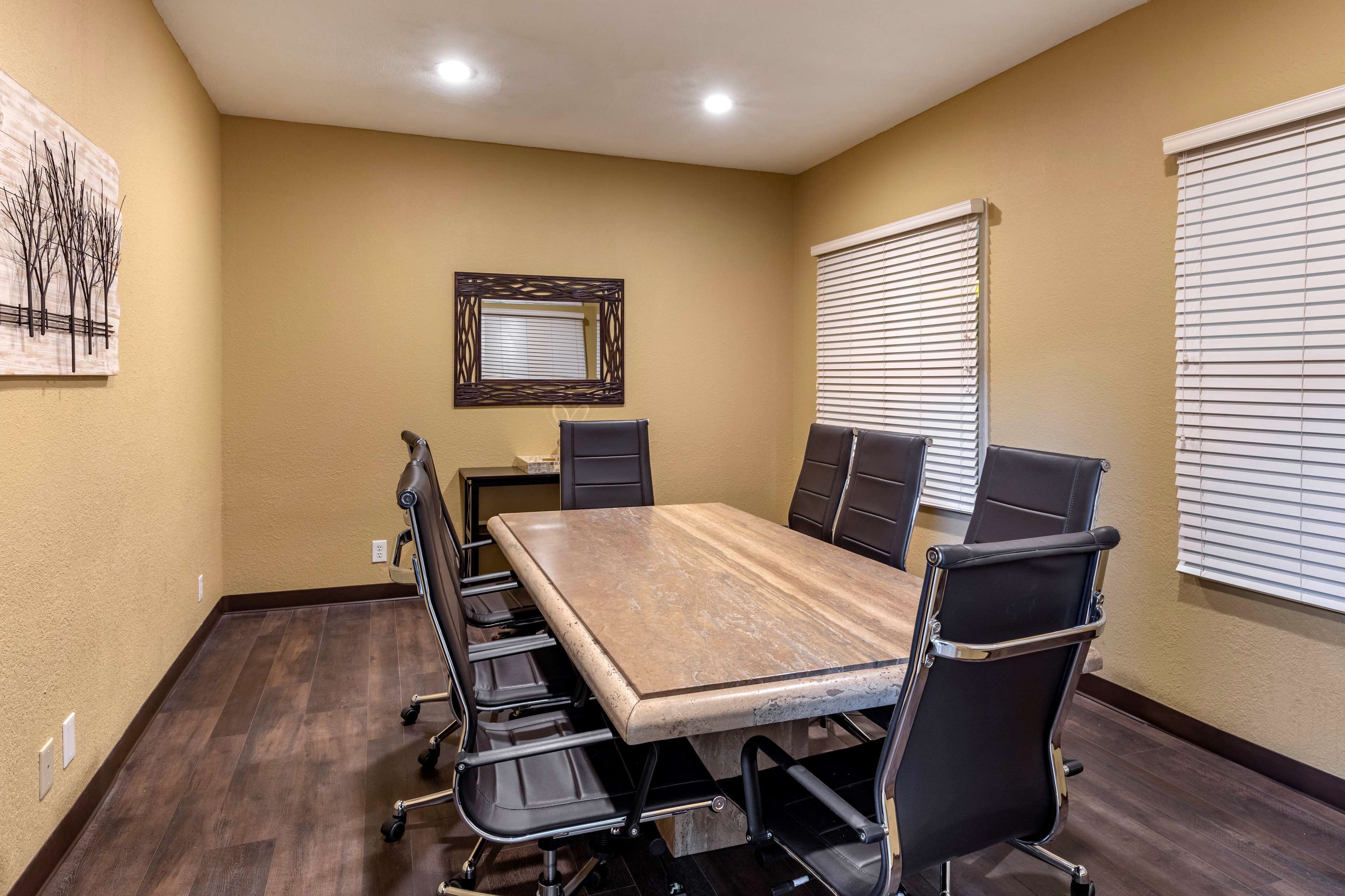 A conference room with a long table, black chairs, a framed mirror, and blinds on the windows.