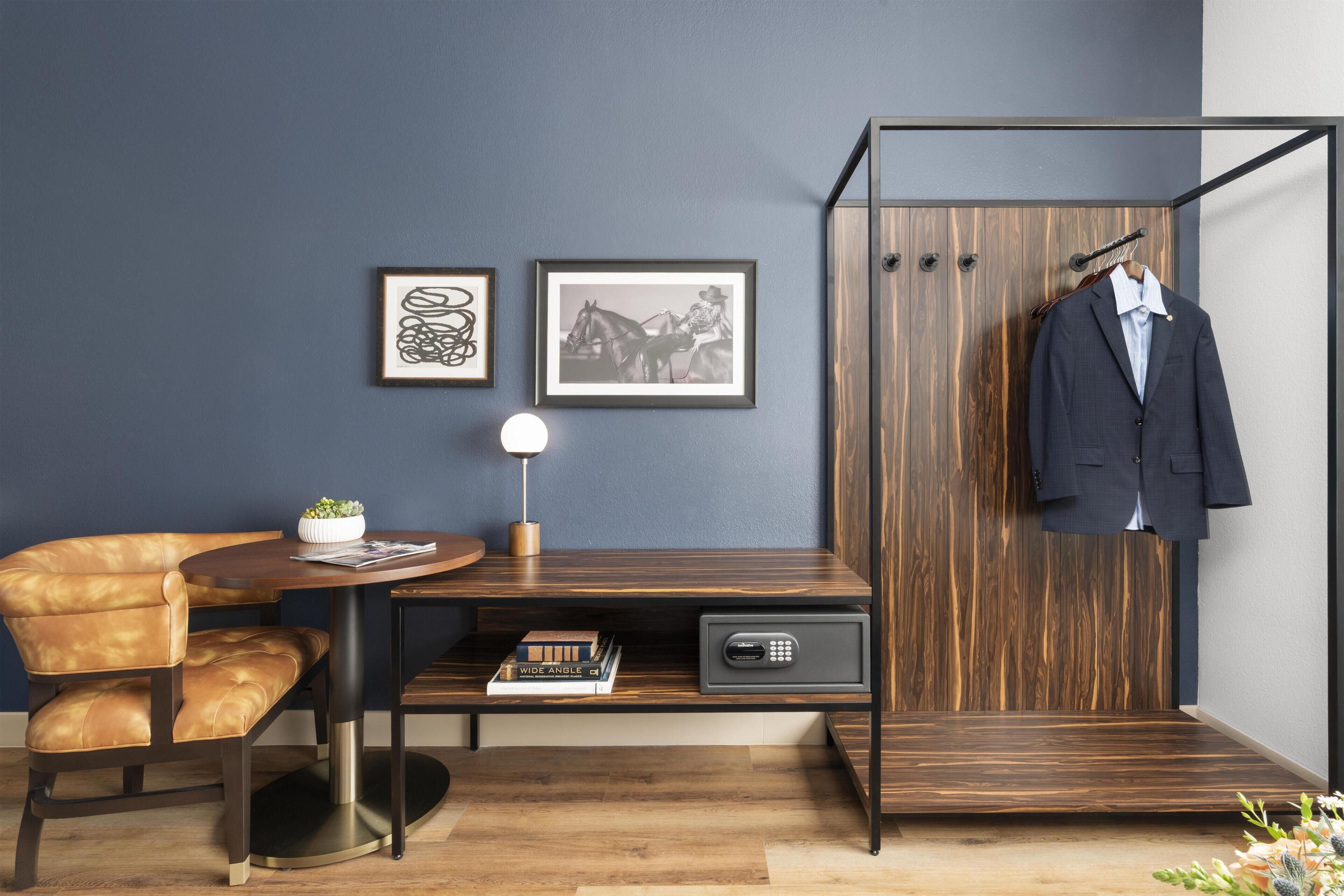 A modern room with a dark blue wall, a wooden desk with a lamp, a leather chair, a coat rack with a suit, and two framed pictures.