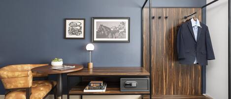 A modern room with a dark blue wall, a wooden desk with a lamp, a leather chair, a coat rack with a suit, and two framed pictures.