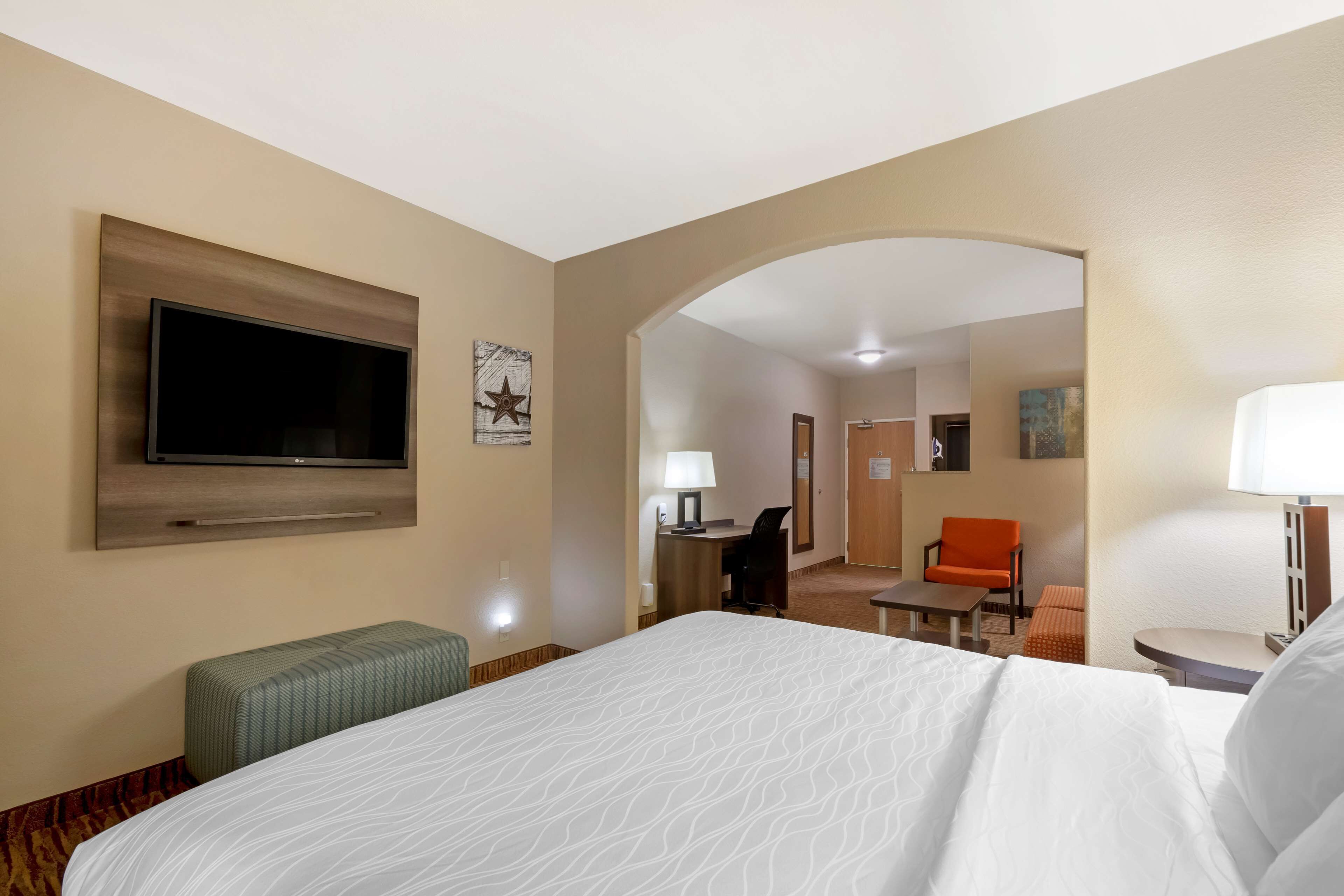 A hotel room with a large bed, a flat-screen TV mounted on the wall, a desk with a chair, and an orange chair.
