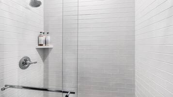 Un baño moderno con azulejos blancos tipo metro, un cabezal de ducha, una ducha de mano, cerramiento de vidrio para la ducha y un estante con artículos de aseo.