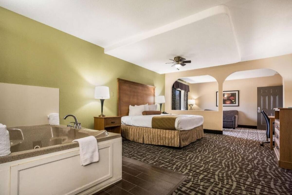 Een hotelkamer met een groot bad, een bed met een houten hoofdeinde, een bureau met een stoel en een plafondventilator.