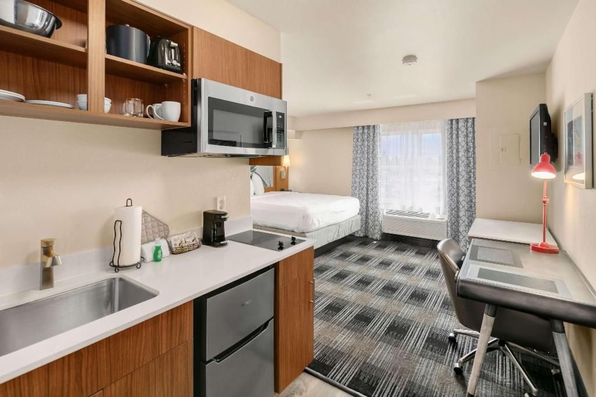 A compact hotel room with a kitchenette, a bed, a desk, and a small bathroom.
