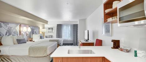 A hotel room with a bed, a small kitchenette, a TV, and a bathroom visible through a glass door.