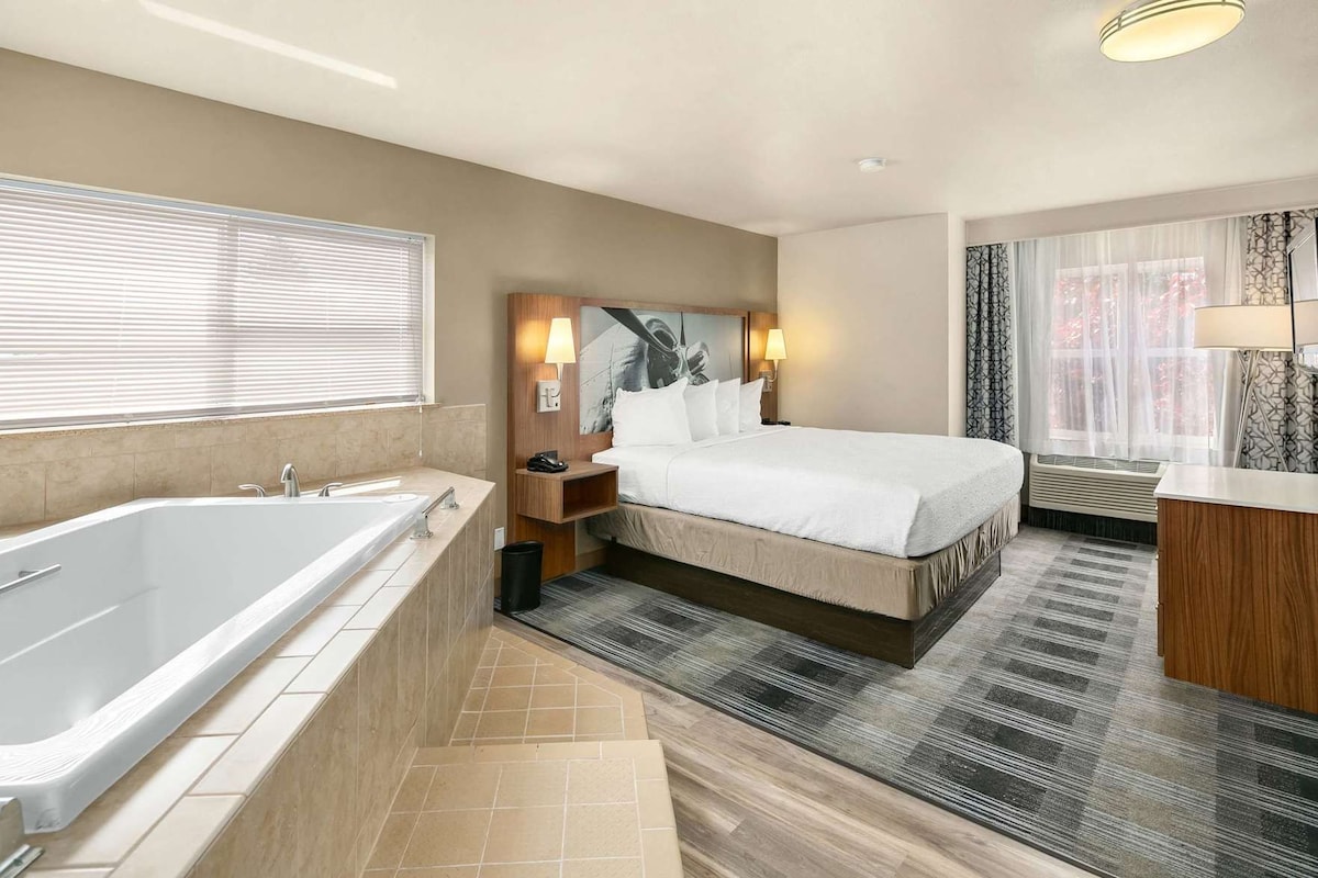 A hotel room with a large bathtub, a bed with a headboard, a bedside table with a lamp, and a window with blinds.