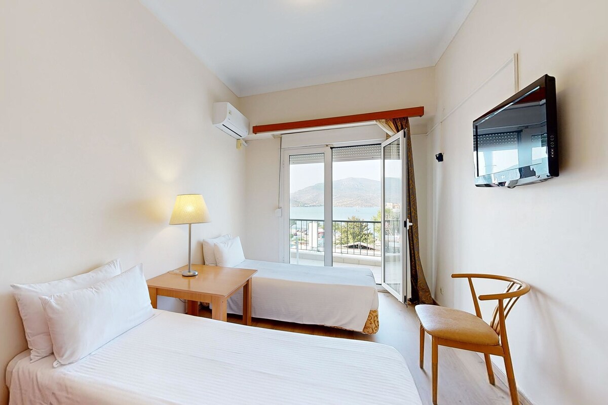 A hotel room with two beds, a desk, a chair, a TV, and a balcony with a view.