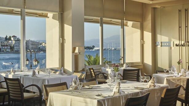 A dining area with tables set for service, large windows offering a view of the sea and buildings, and a modern interior design. - Lucy Hotel (Chalcis)