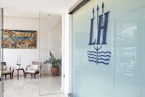 A modern interior with a glass wall displaying a logo, a seating area with chairs and a table, and a large abstract painting on the wall. - Lucy Hotel (Chalcis)