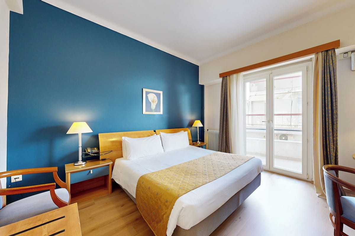 A hotel room with a large bed, two bedside tables with lamps, a desk with a chair, and a window with curtains.