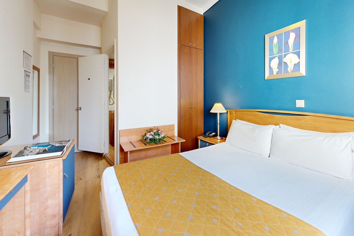 A hotel room with a bed, a desk with a computer, a nightstand with a lamp, and a wall with a framed picture.