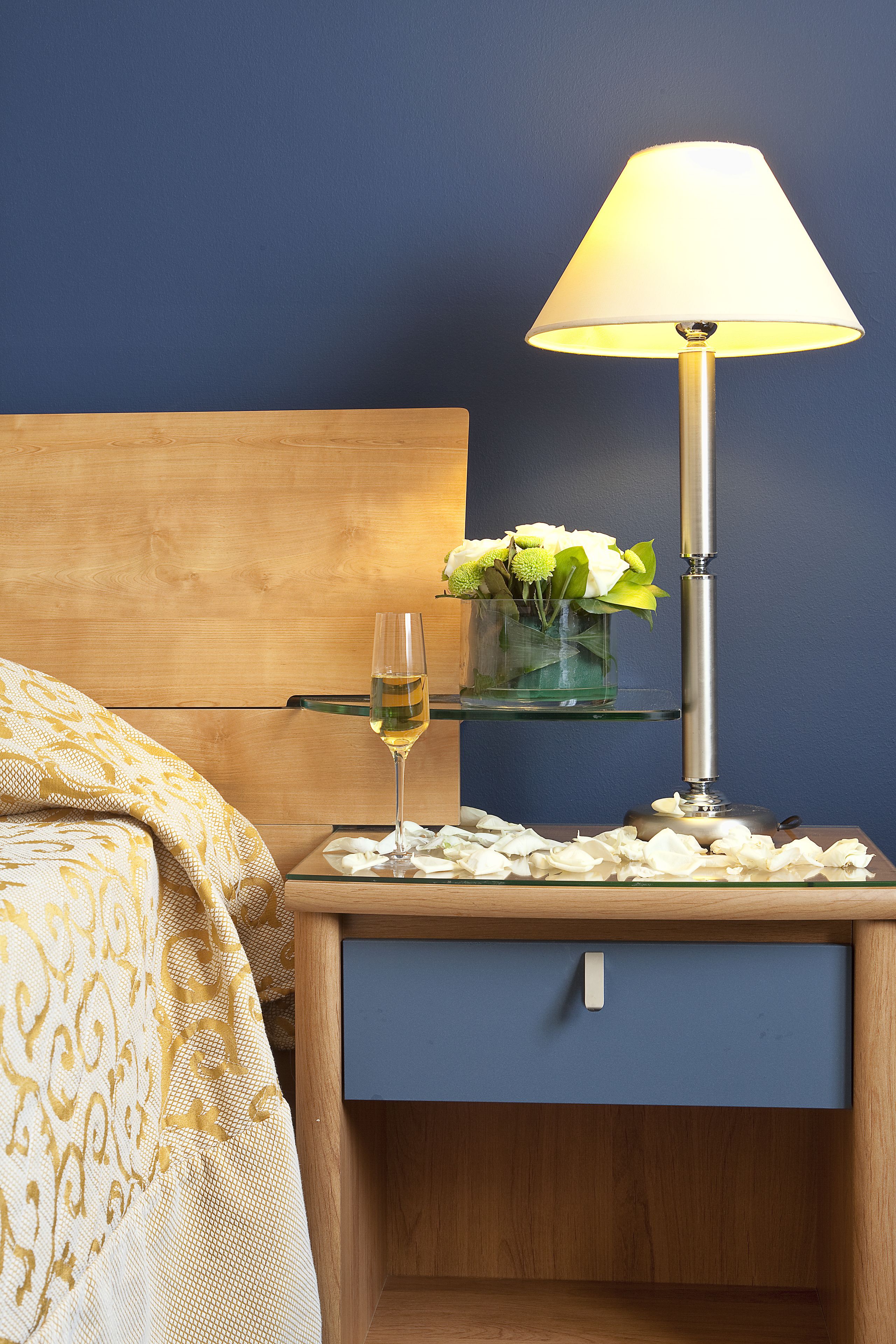 A neatly made bed with a patterned blanket, a bedside table with a glass vase of flowers, a glass of white wine, and a lamp.
