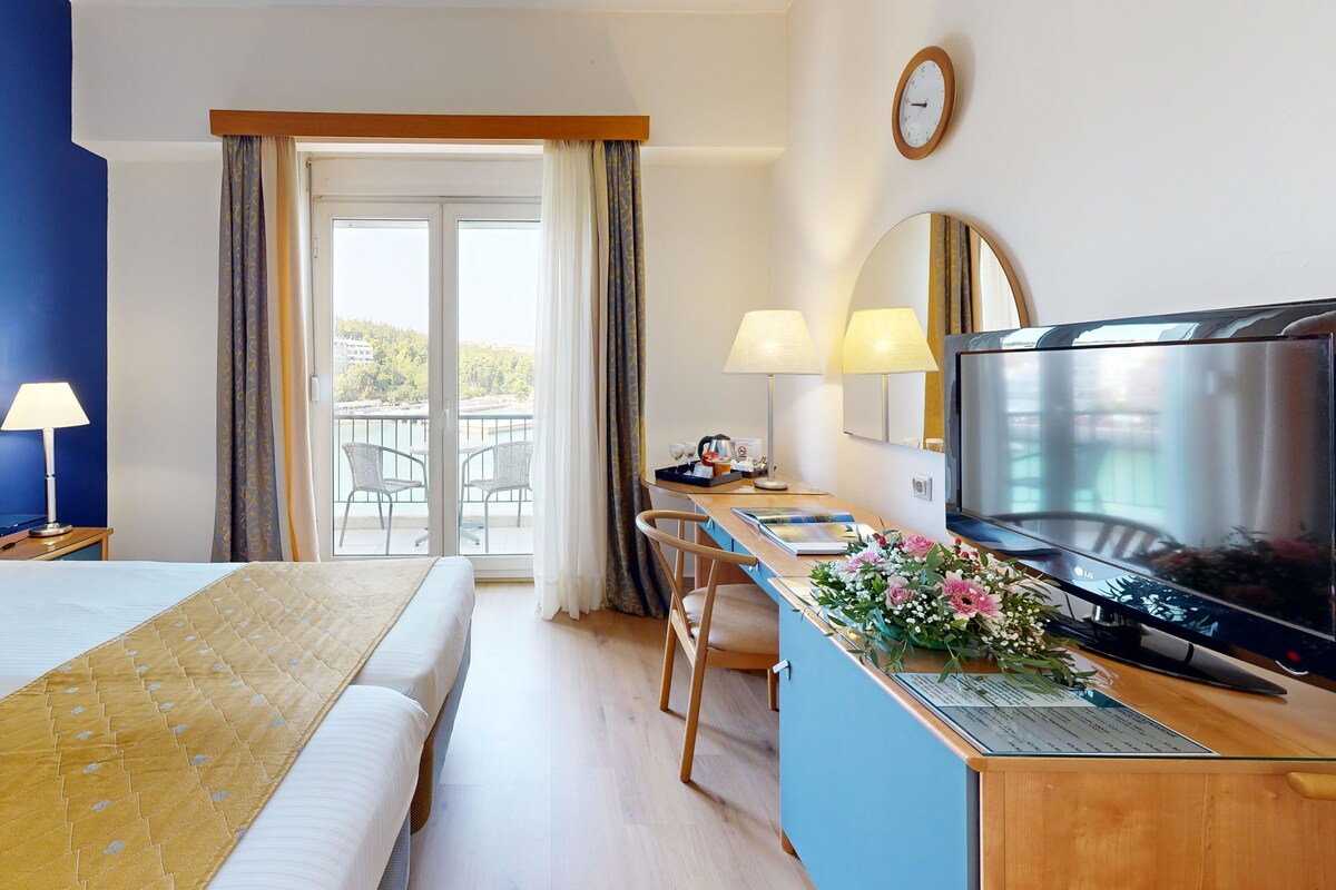 A hotel room with a bed, a desk with a TV, a chair, and a balcony with a view.