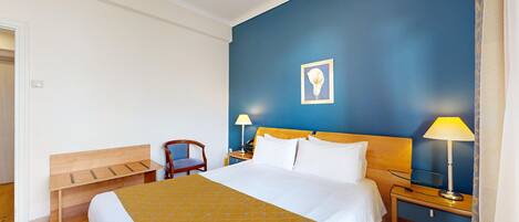 Habitación de hotel con cama, un banco de madera, una silla, dos mesitas de noche con lámparas y un cuadro enmarcado en la pared.