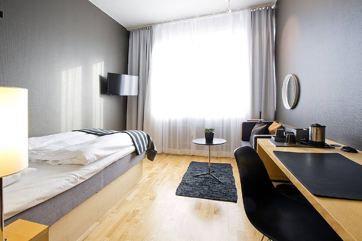 Een moderne hotelkamer met een enkel bed, een bureau, een stoel, een klein tafeltje, een televisie en een raam met gordijnen.
