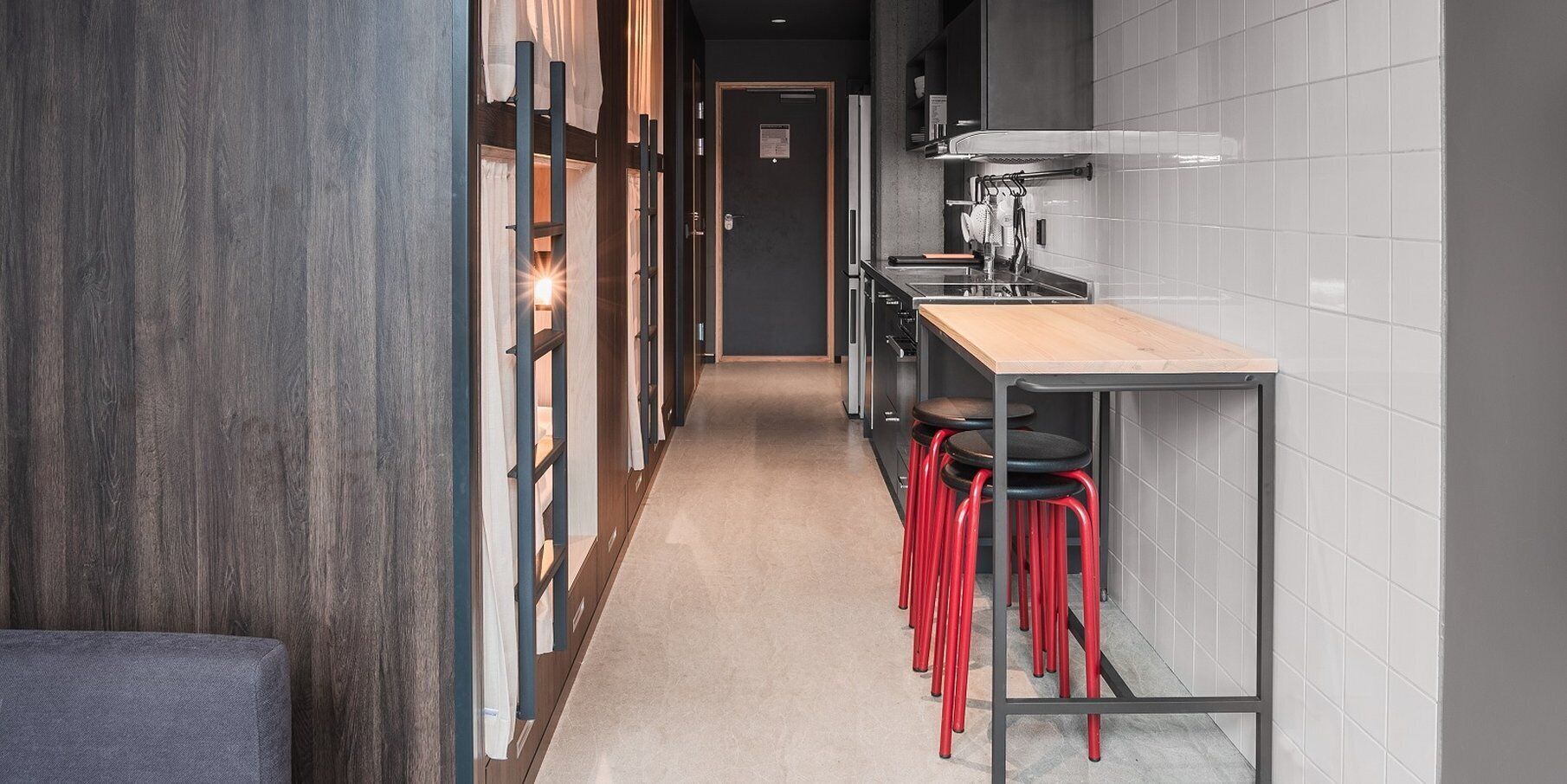 A narrow hallway with a wooden floor, a long table with red stools, and a kitchen area with white tiles and a stove.