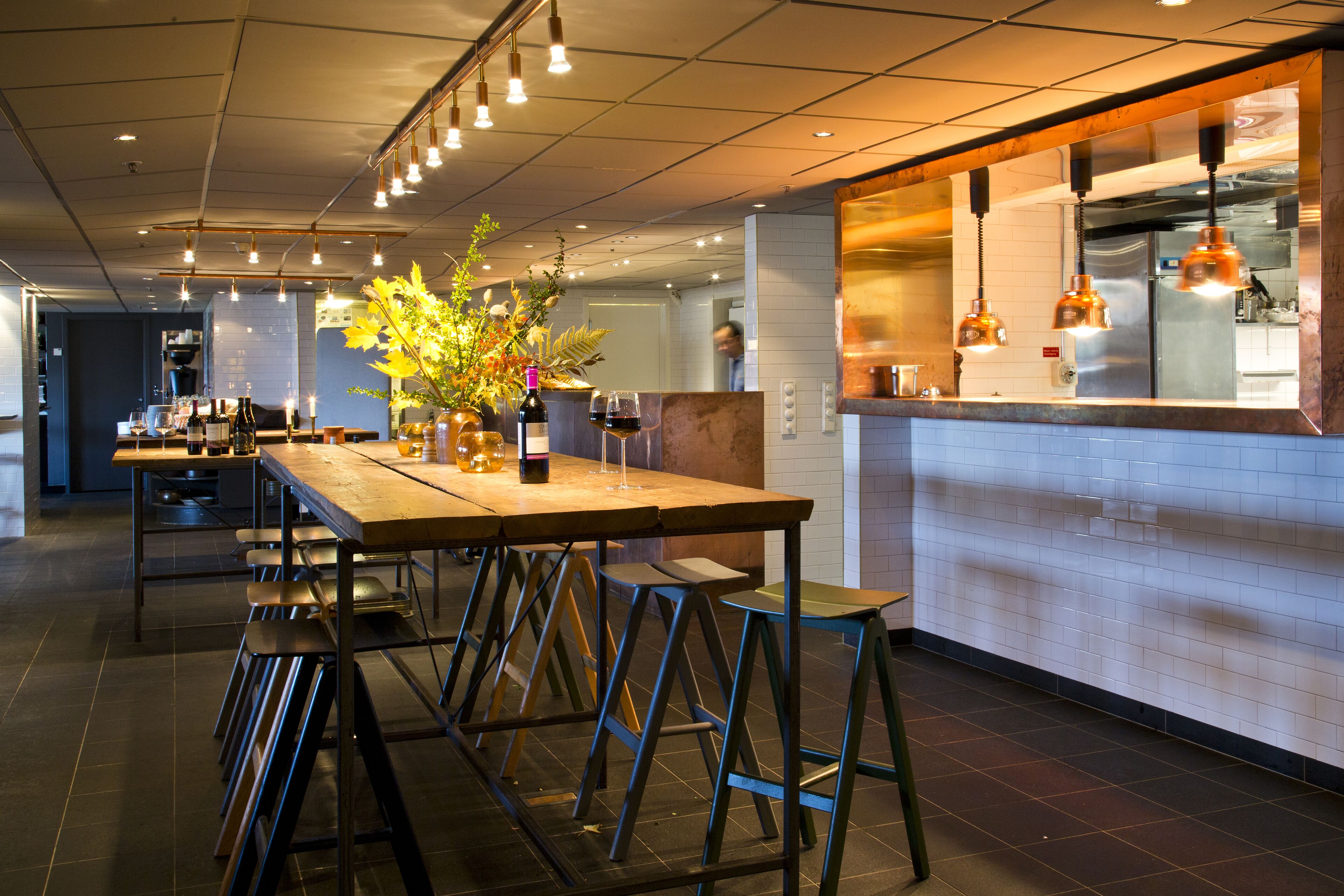 Un interior de restaurante moderno con una larga mesa de madera, taburetes de bar, lámparas colgantes y una zona de cocina visible a través de una partición de vidrio.
