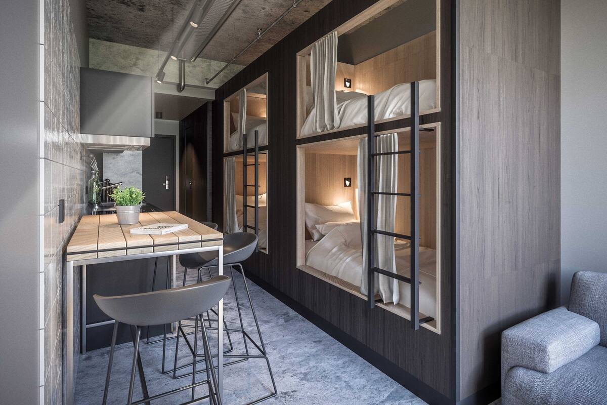 Een compacte hotelkamer met een stapelbed, een kleine kitchenette, een zithoek met een tafel en stoelen, en een groot raam met gordijnen.