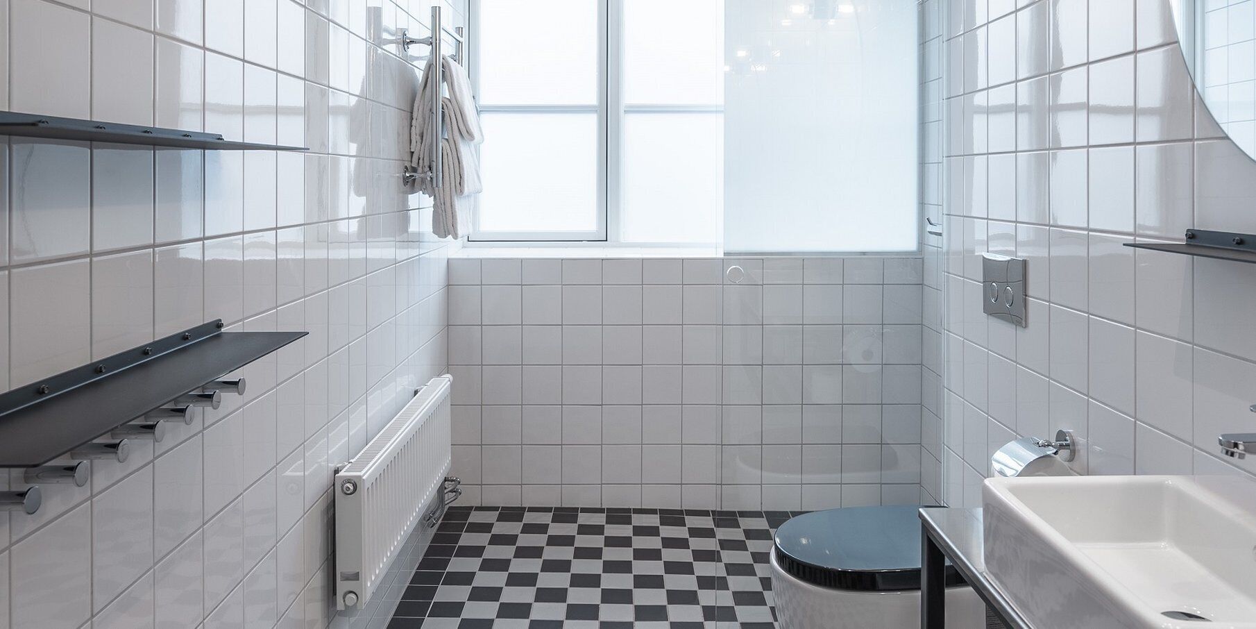 A small, modern bathroom with white tiles, a black and white checkered floor, a window, a radiator, and a toilet.