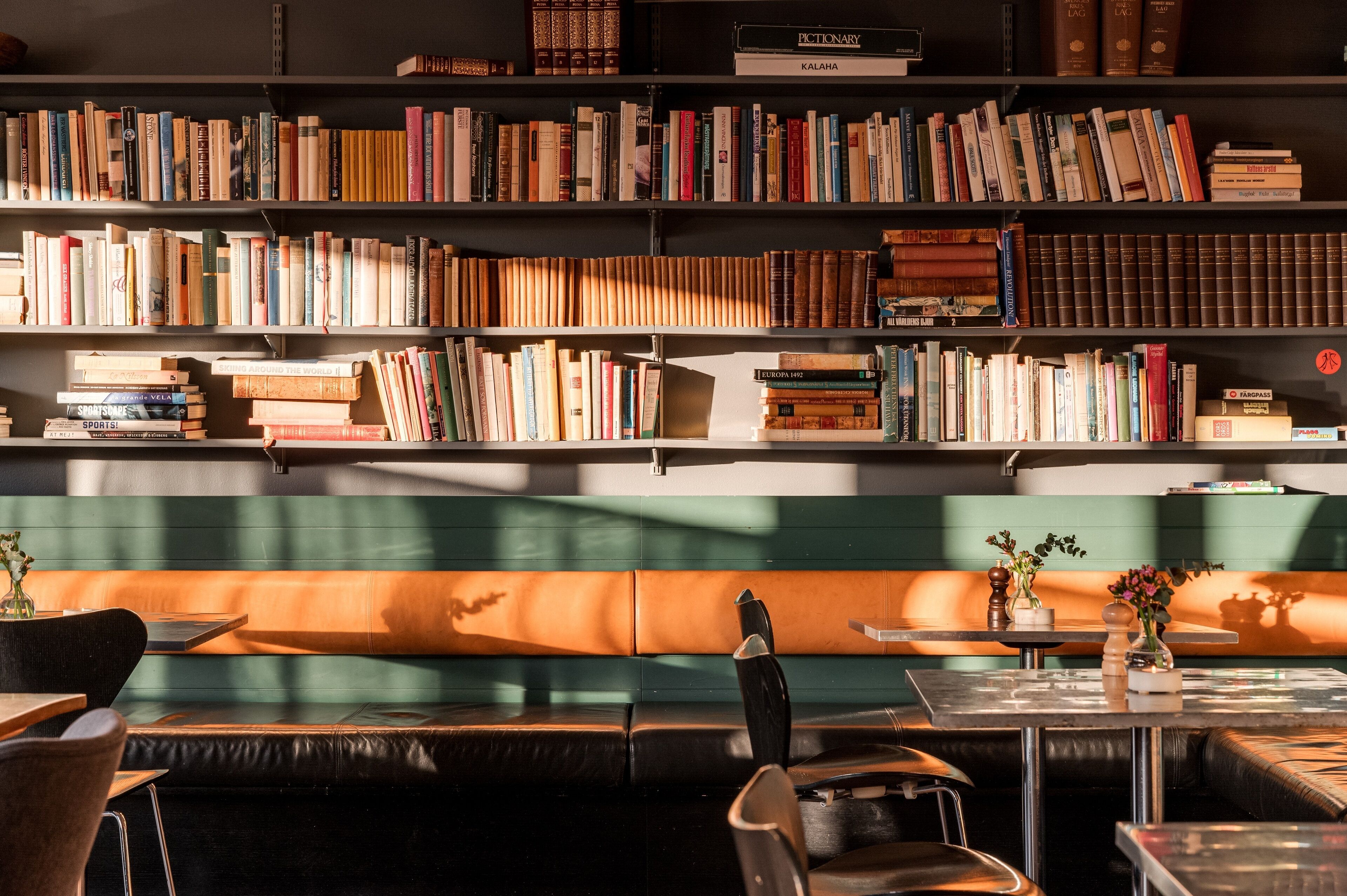 Una biblioteca con estanterías repletas de libros, un área de descanso con un banco y mesas, y un pequeño jarrón con flores sobre una de las mesas.