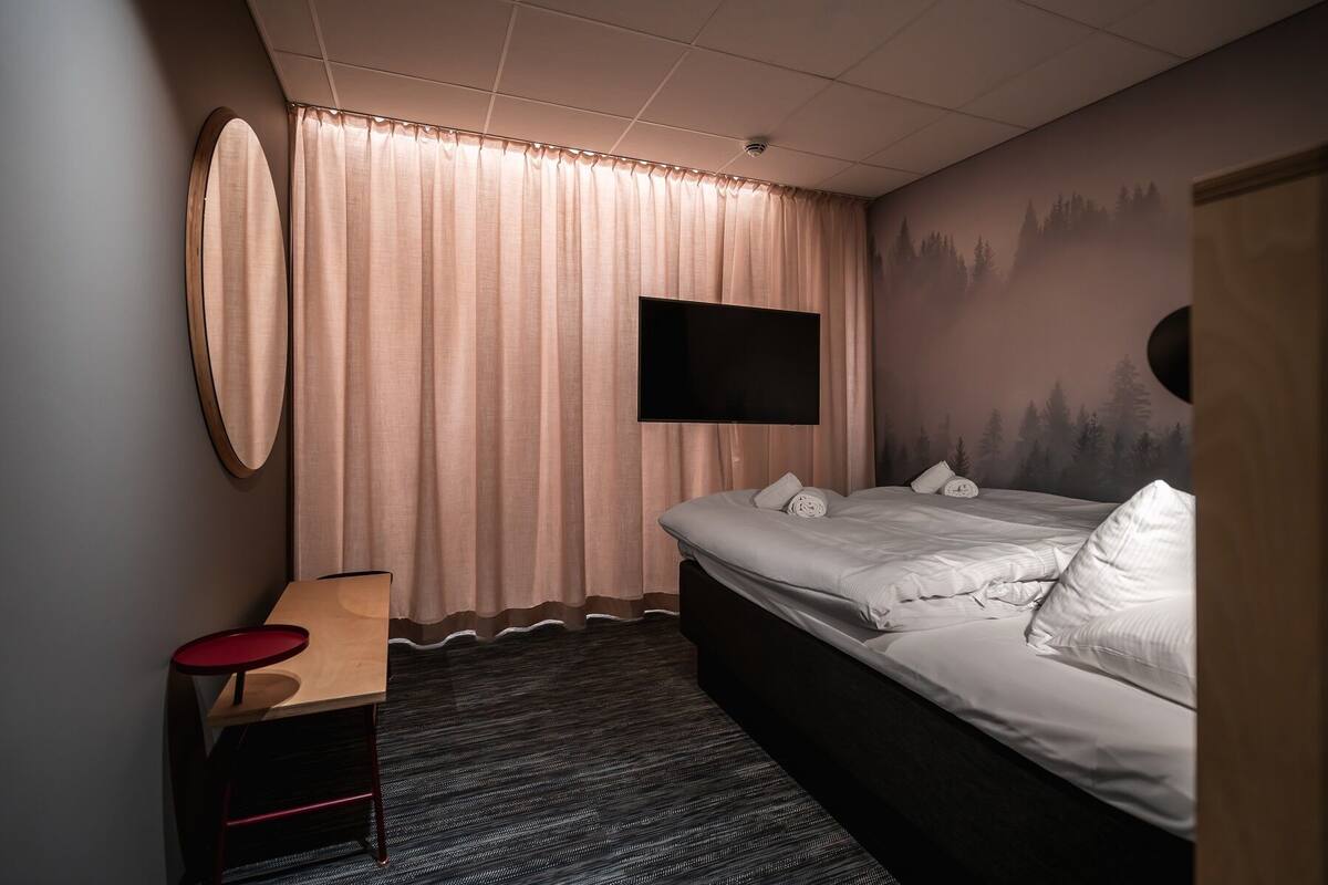 Een hotelkamer met een bed, een aan de muur bevestigde televisie, een bureau met stoel en een groot raam met gordijnen.