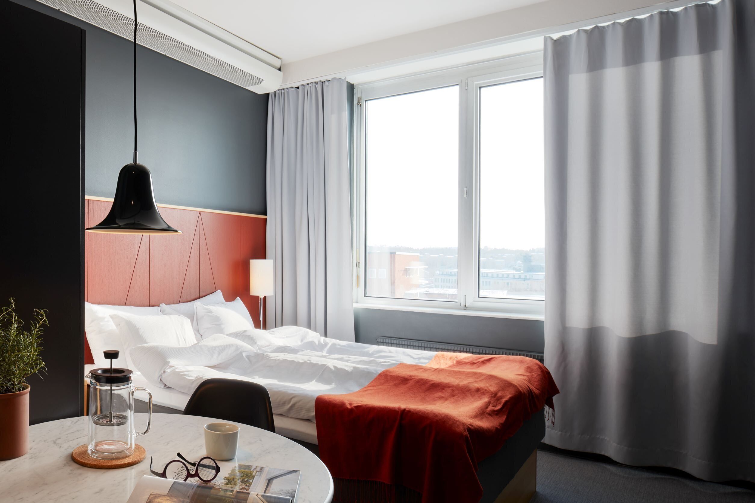 Una camera d'albergo con un letto, una macchina del caffè, un tavolino con una tazza e dei bicchieri, e una finestra con tende.