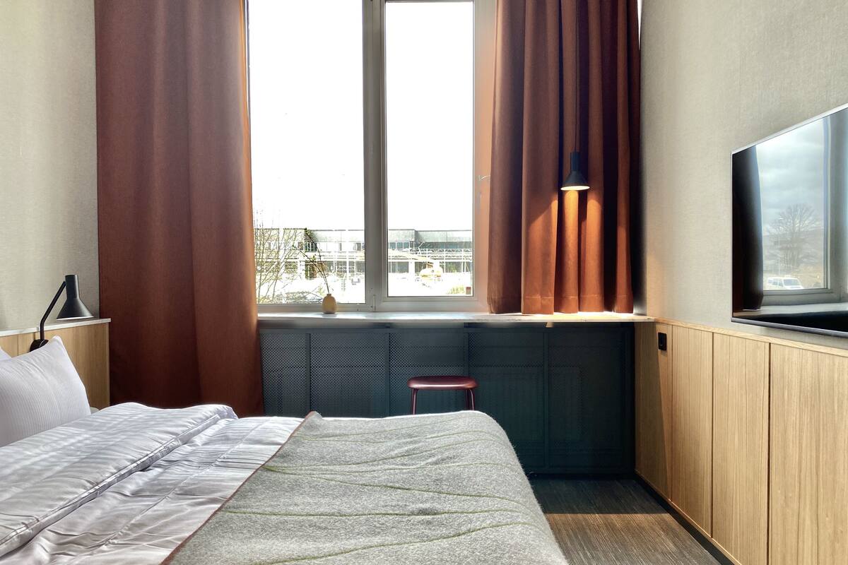 Een hotelkamer met een bed, een raam met gordijnen, een radiator, een klein krukje en een televisie aan de muur.