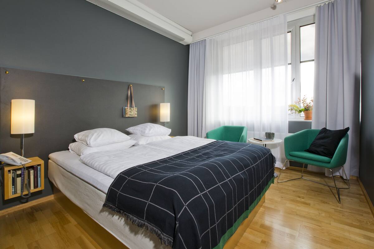 Een moderne hotelkamer met een groot bed, bedlampen, een groene fauteuil en een klein tafeltje met een bloempot.