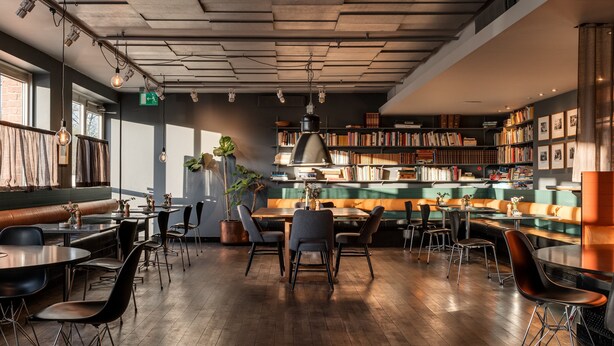 Un café moderno con suelos de madera, asientos negros y marrones, y una gran estantería llena de libros.