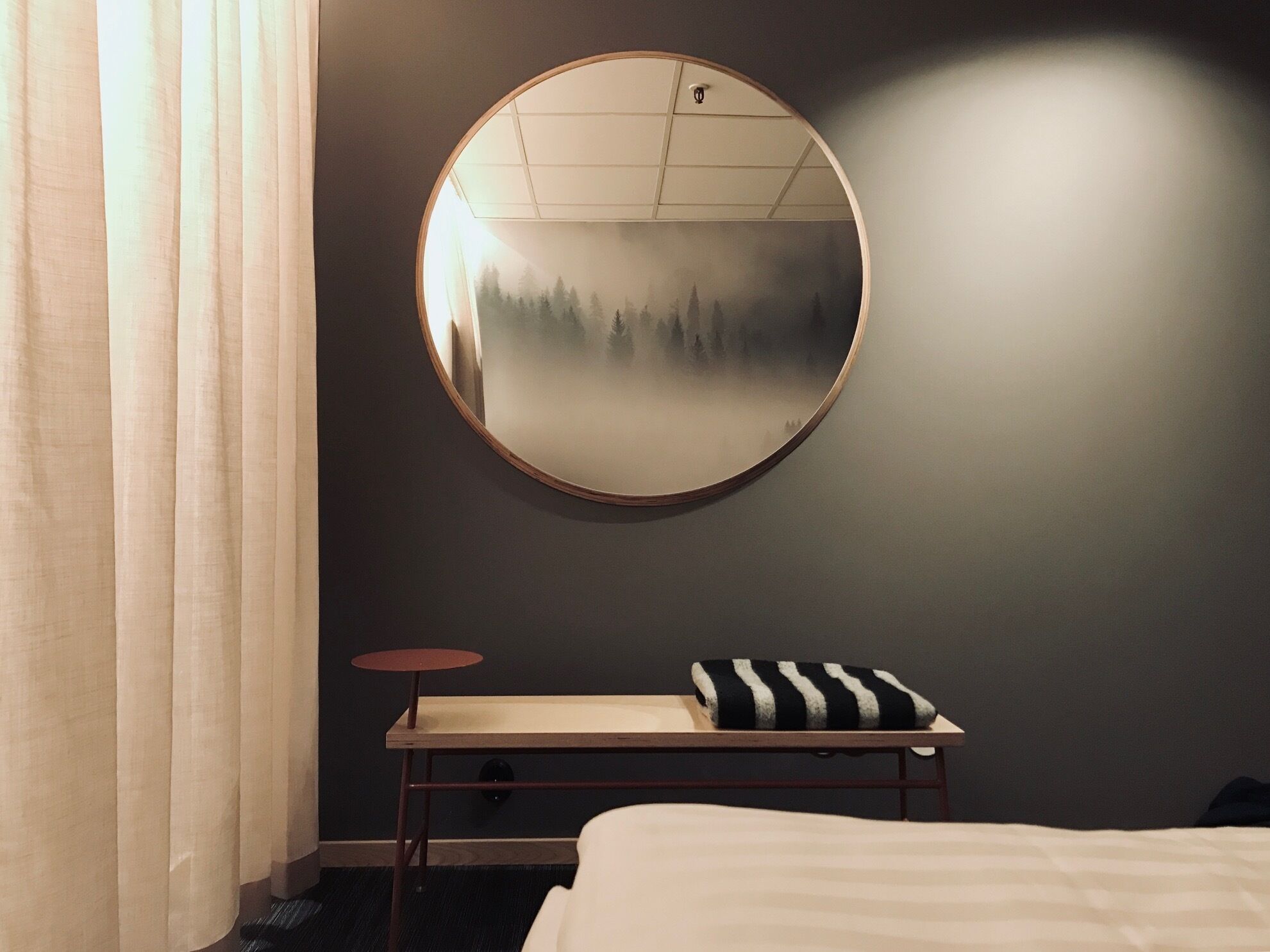 Una habitación de hotel con un espejo redondo en la pared, una mesita con un cojín a rayas y una cama con edredón blanco.