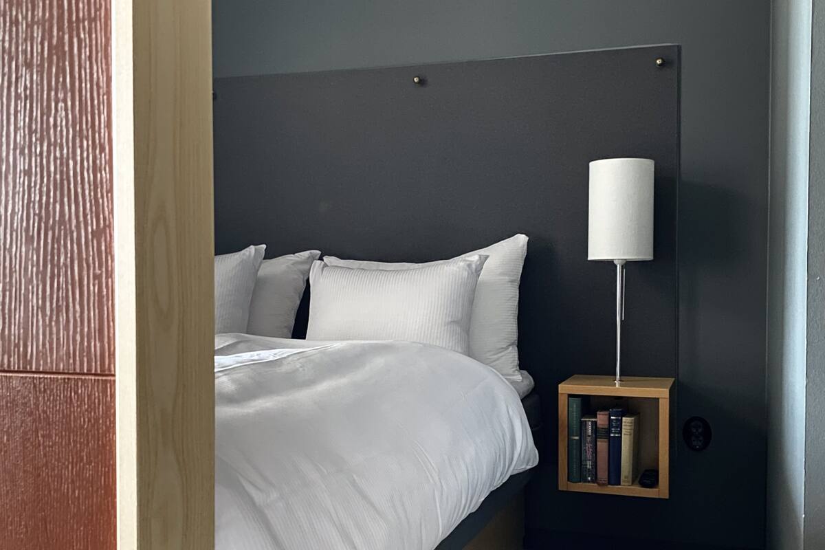 Een net opgemaakt bed met witte lakens, een bedlampje en een klein houten nachtkastje met boeken.