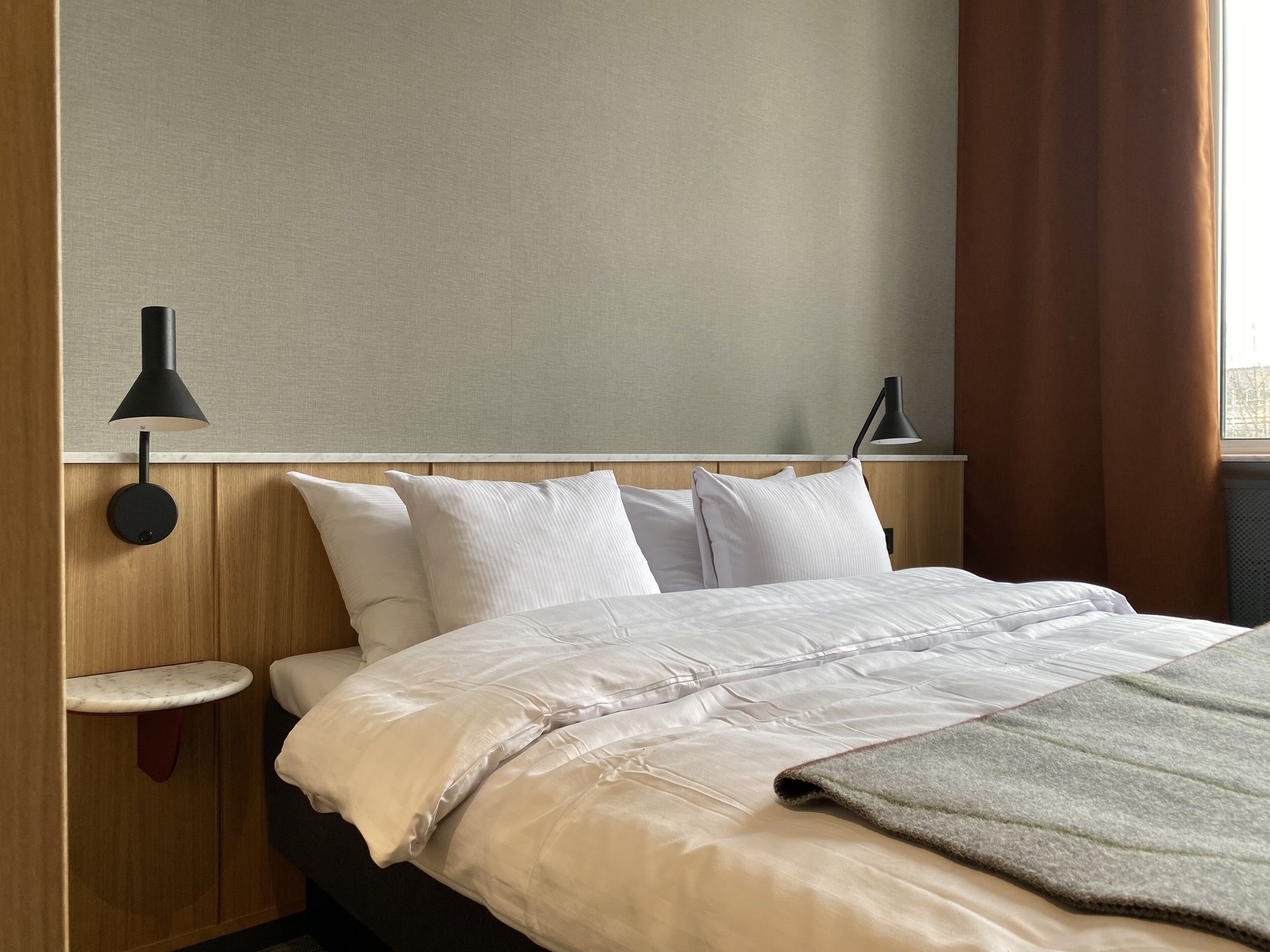 A hotel room with a bed, two bedside tables, a wall-mounted lamp, and a window with curtains.