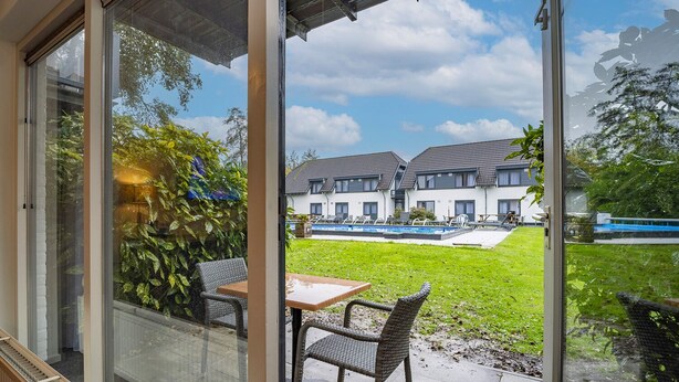 A glass door leading to a patio with wicker furniture, a pool, and a row of houses in the background. - Fletcher Hotel De Witte Brug (Lekkerkerk)