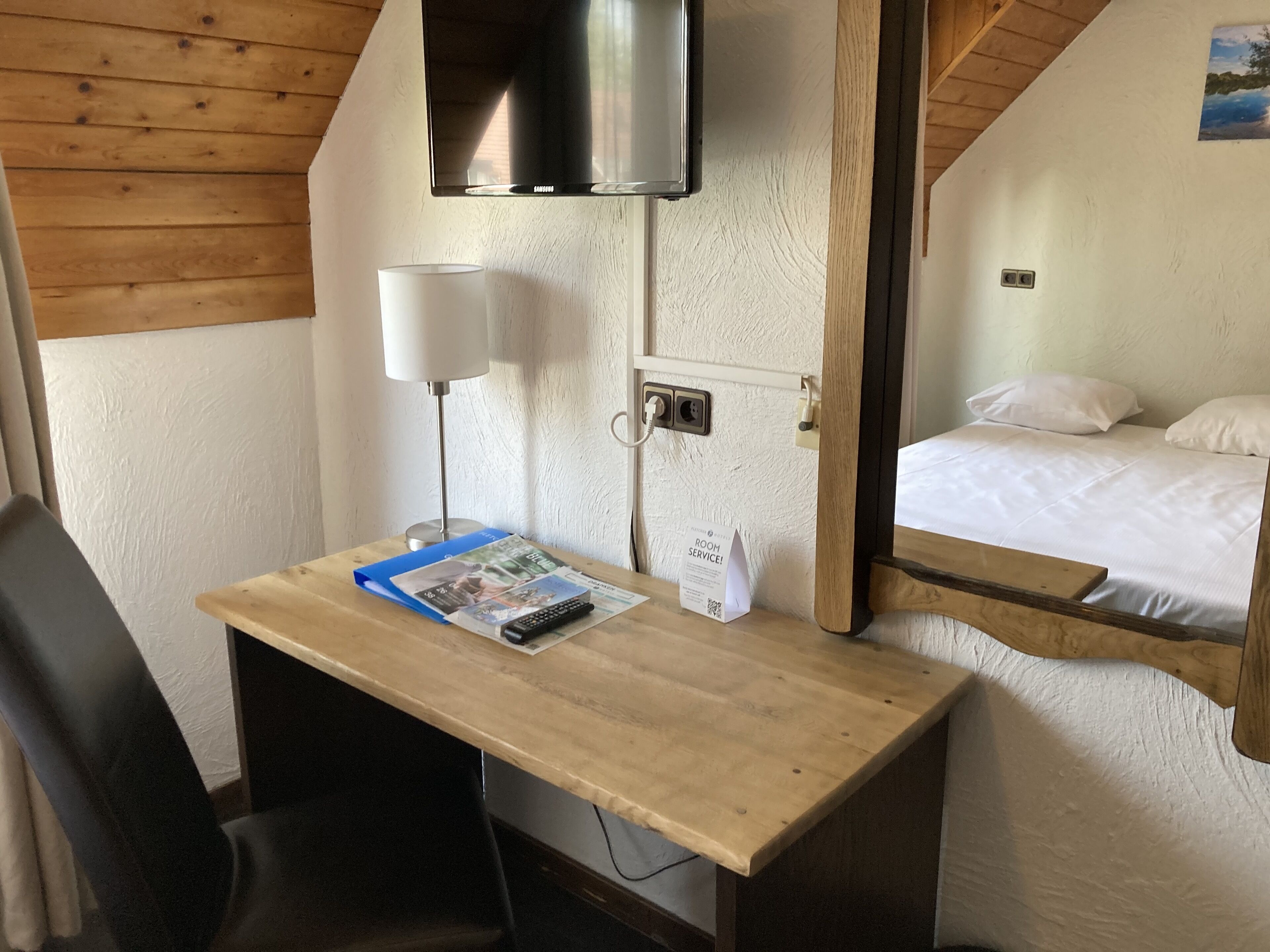 Una habitación de hotel con un escritorio de madera, una lámpara, un televisor montado en la pared y una cama con ropa blanca.