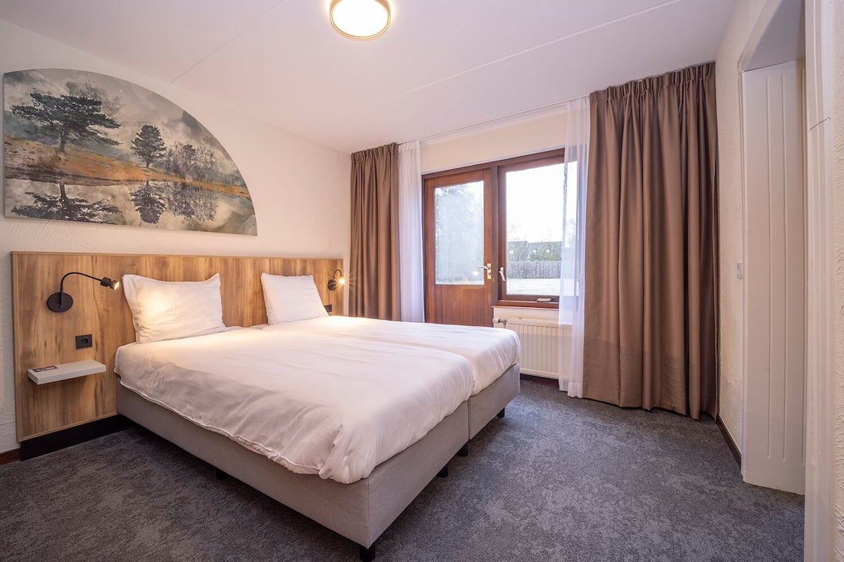 A hotel room with a large bed, a headboard with a light fixture, a painting on the wall, a window with curtains, and a radiator.
