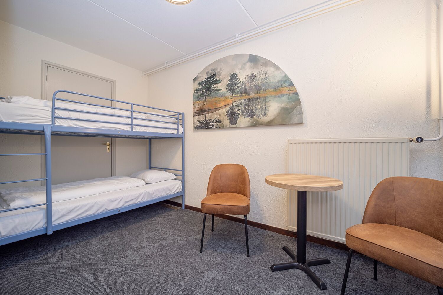 A bunk room with two beds, a round table, two chairs, and a painting on the wall.
