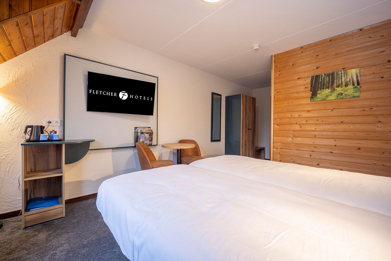 Habitación de hotel con cama, escritorio con tetera, silla, estantería y un cuadro en la pared.