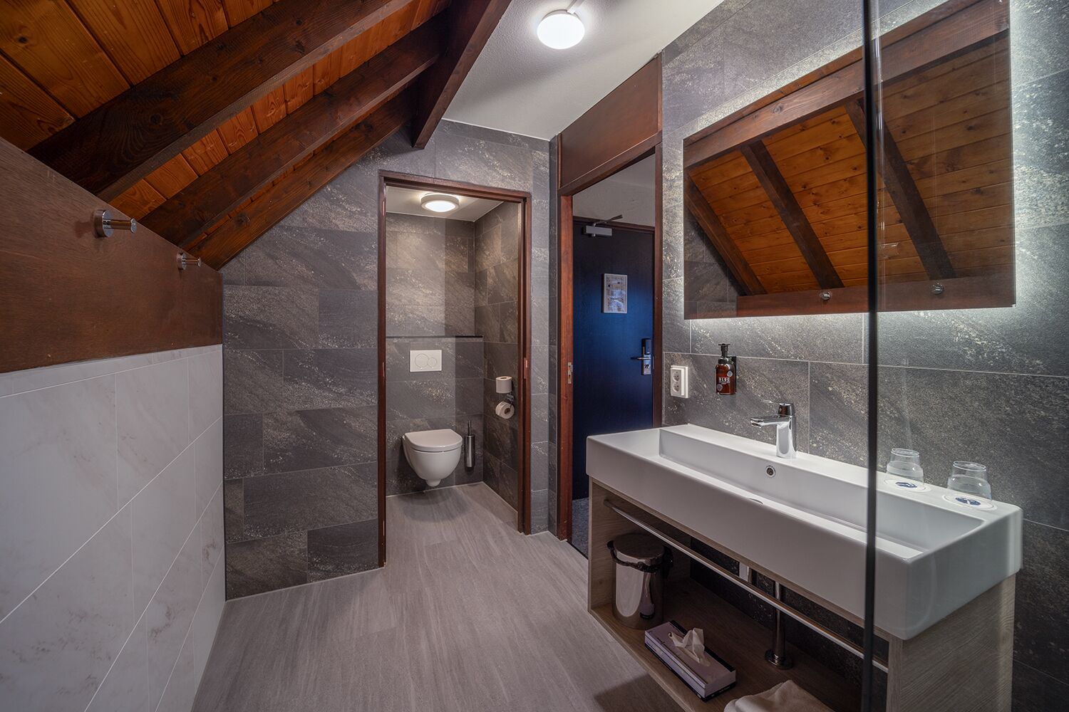 A modern bathroom with a shower, toilet, and sink, featuring stone tiles and wooden beams.