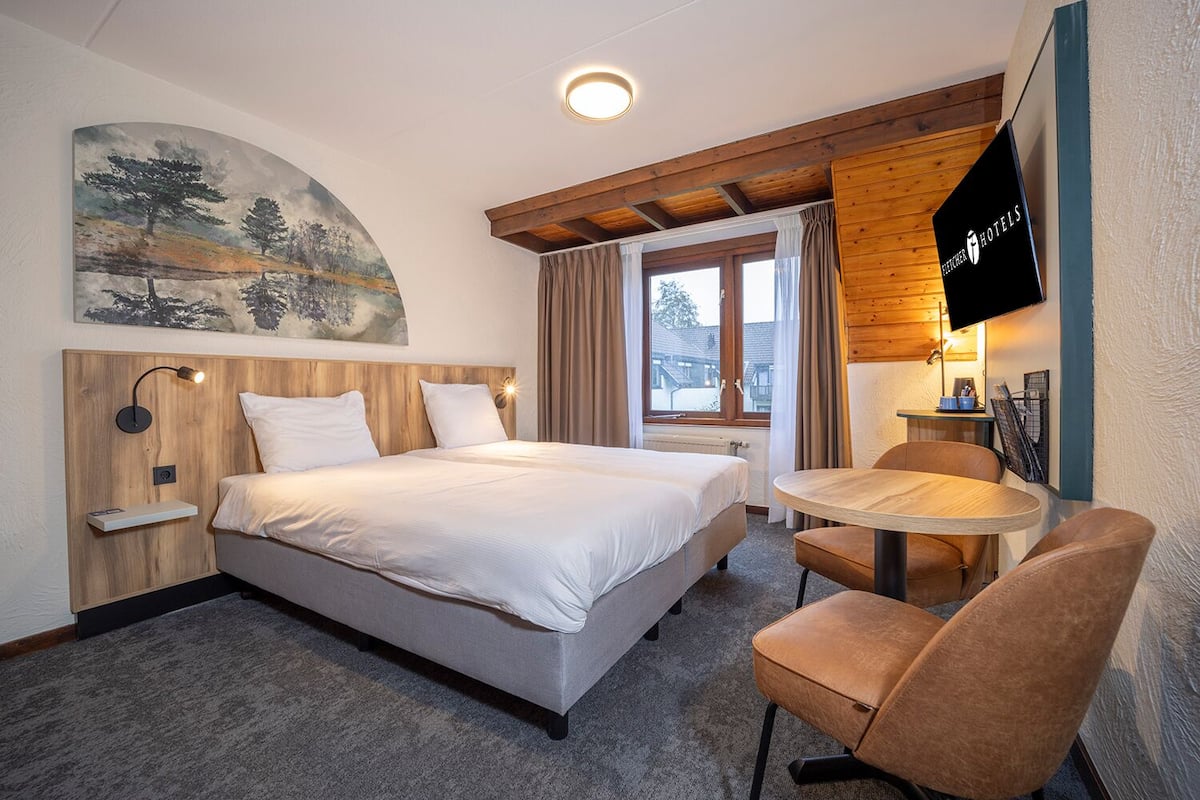 A hotel room with a bed, a desk, a chair, a TV, and a painting on the wall.