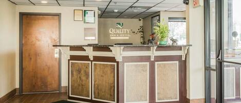 A reception area with a wooden door, a reception desk with a sign reading "Quality Inn & Suites," and a green doormat.