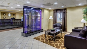 A hotel lobby with a reception desk, a seating area with a sofa and armchair, a decorative glass partition, and a floor lamp.