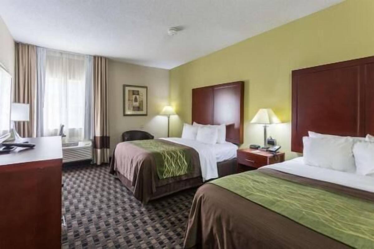 A hotel room with two beds, a desk, a chair, and a lamp.