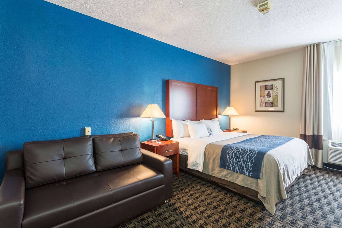 A hotel room with a blue accent wall, a bed with a wooden headboard, a brown leather sofa, two bedside lamps, and a framed picture on the wall.