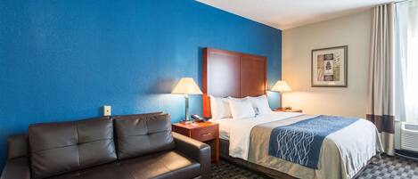 A hotel room with a blue accent wall, a bed with a wooden headboard, a brown leather sofa, two bedside lamps, and a framed picture on the wall.
