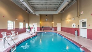 An indoor swimming pool with a tiled edge, a lifeguard chair, and a few white plastic chairs around the pool.