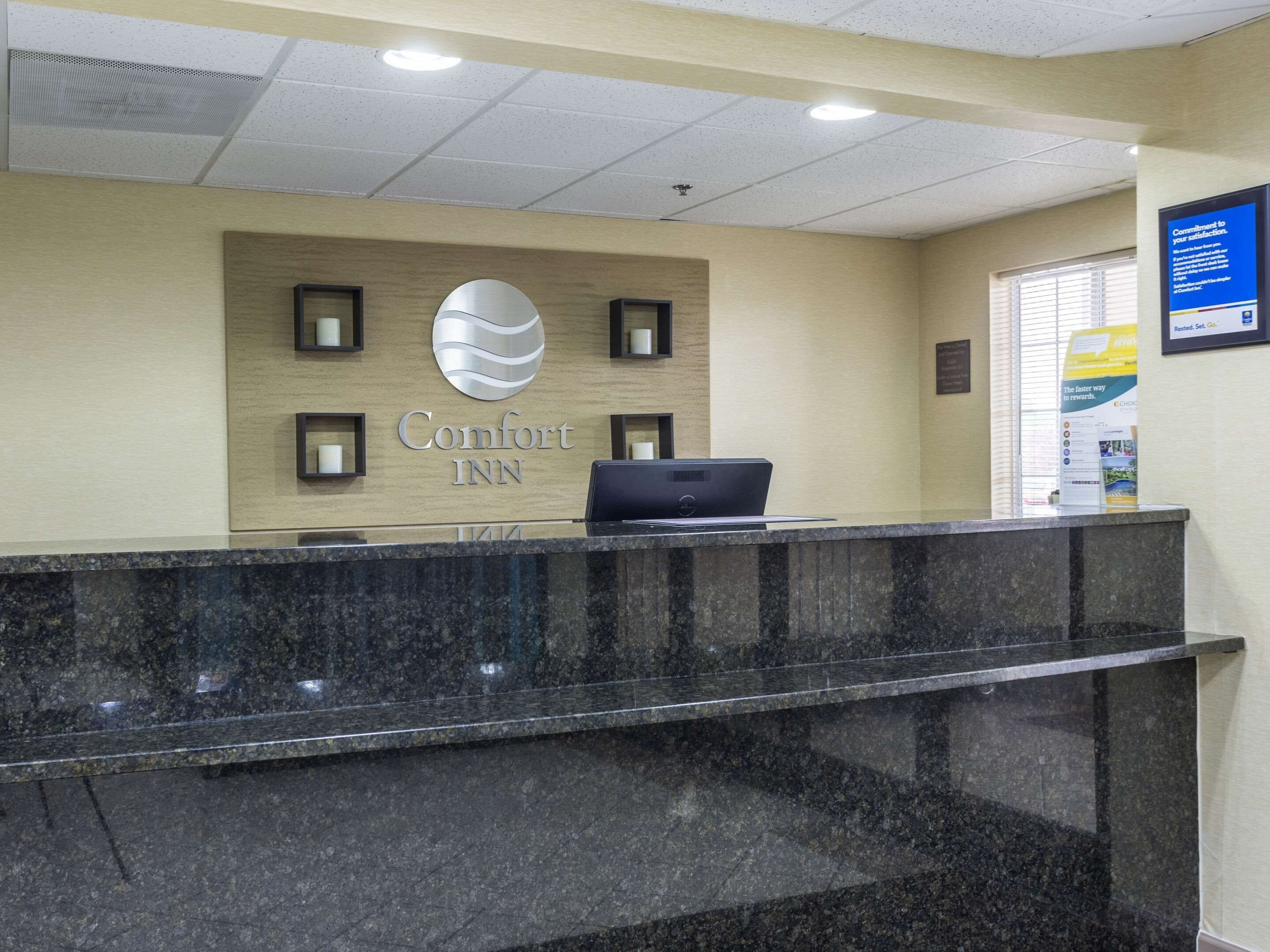 A reception desk at Comfort Inn with a laptop, a large Comfort Inn logo, and a framed sign on the wall.