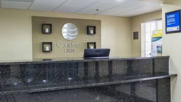 A reception desk at Comfort Inn with a laptop, a large Comfort Inn logo, and a framed sign on the wall.