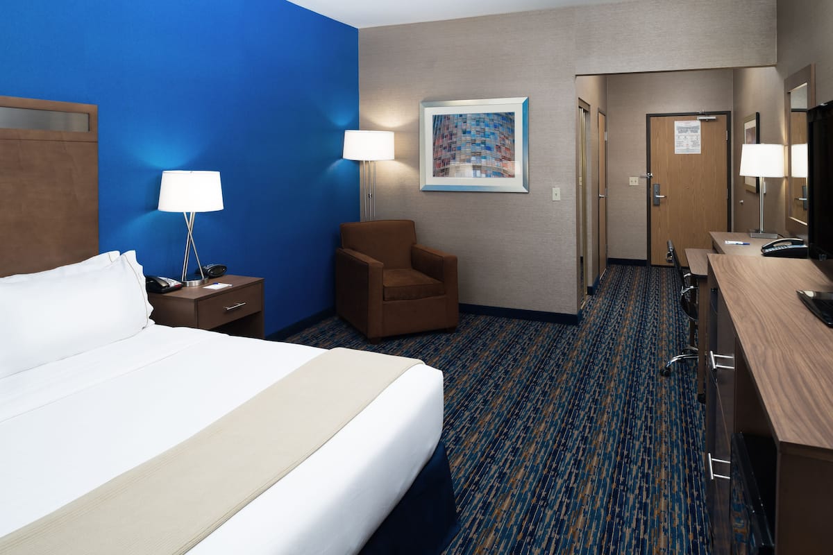 A hotel room with a large bed, a chair, a desk, a lamp, and a painting on the wall.