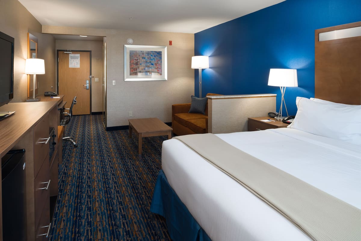 A hotel room with a large bed, a desk with a lamp, a chair, a small table, and a nightstand with a lamp.