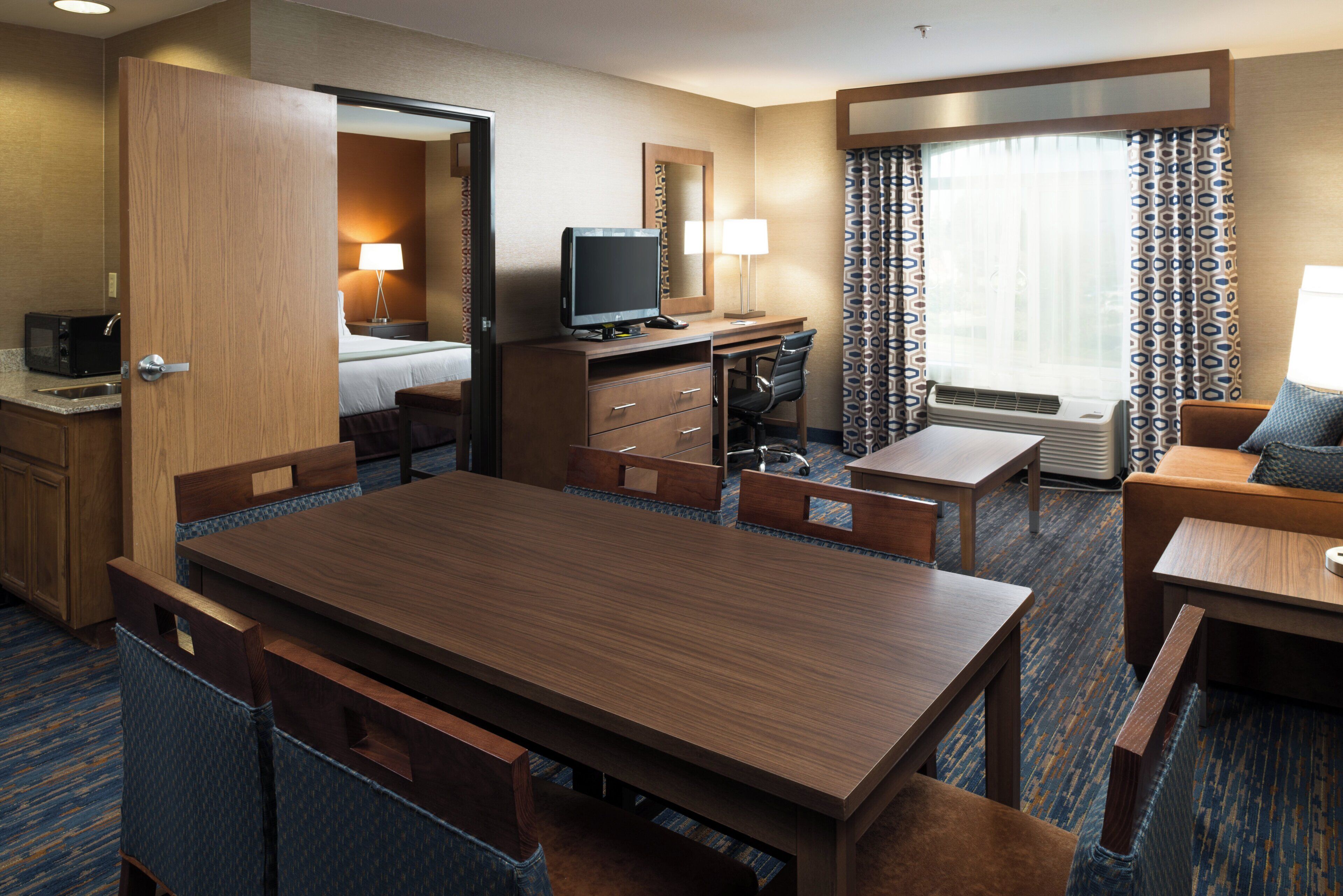 A hotel room with a wooden dining table, a separate desk with a computer, a bed, a bathroom, and a large window with patterned curtains.